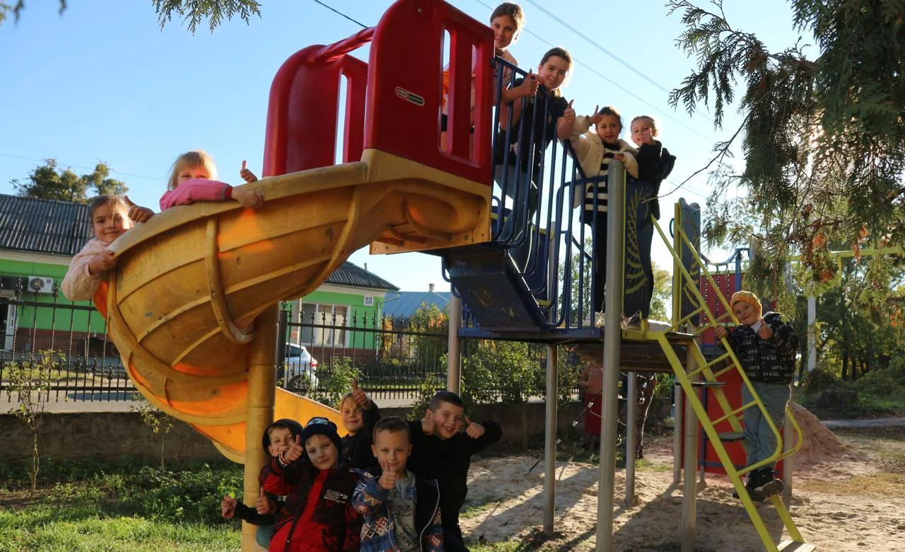 Children on a playground slide and climbing frame, many giving a thumbs-up