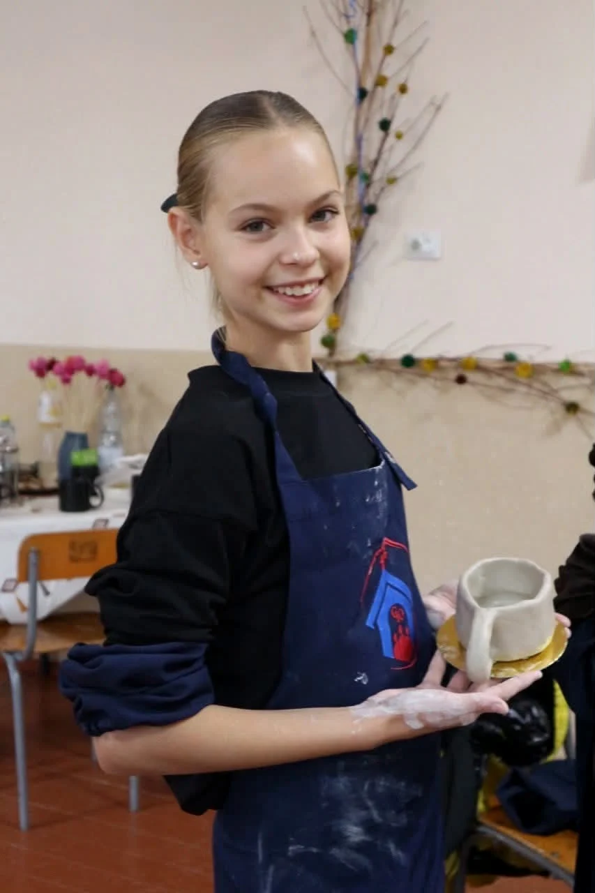 A smiling girl in a studio apron holding a handmade clay cup and saucer
