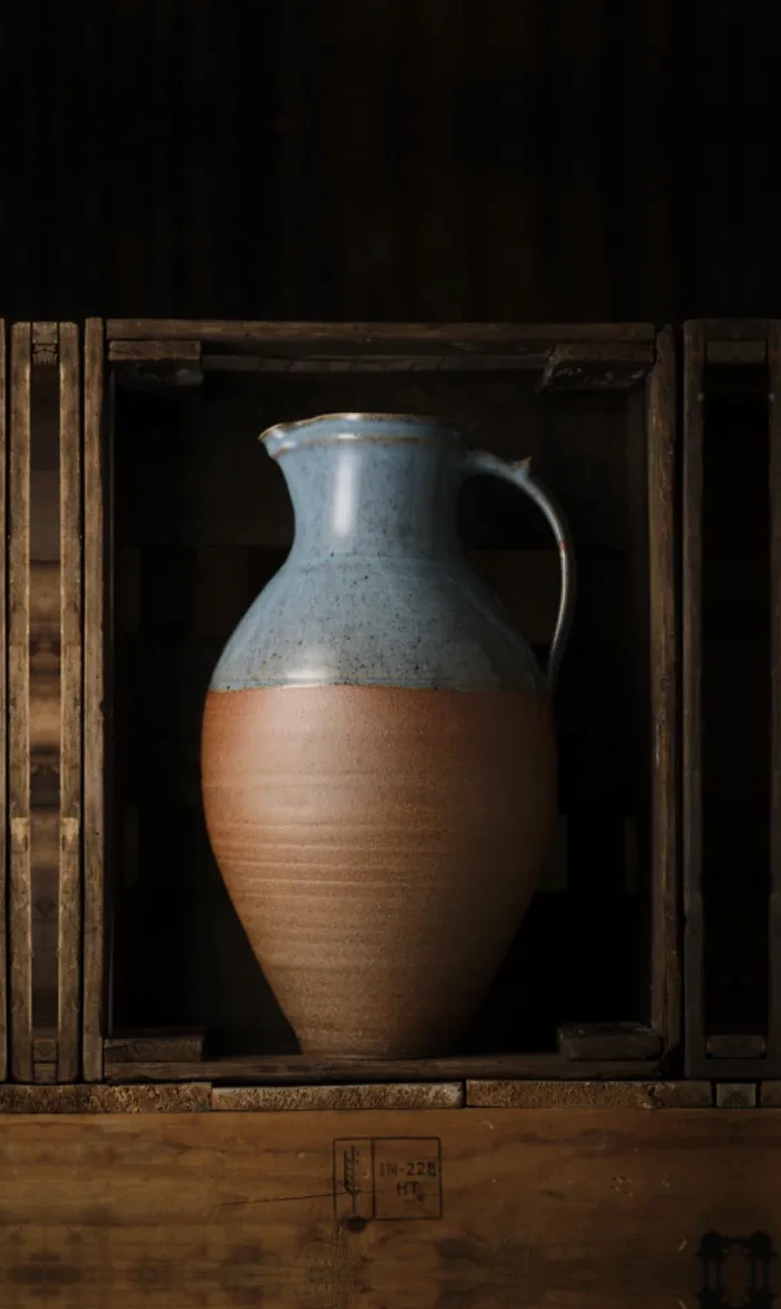 A hand-thrown stoneware jug with a blue glaze in a wooden crate